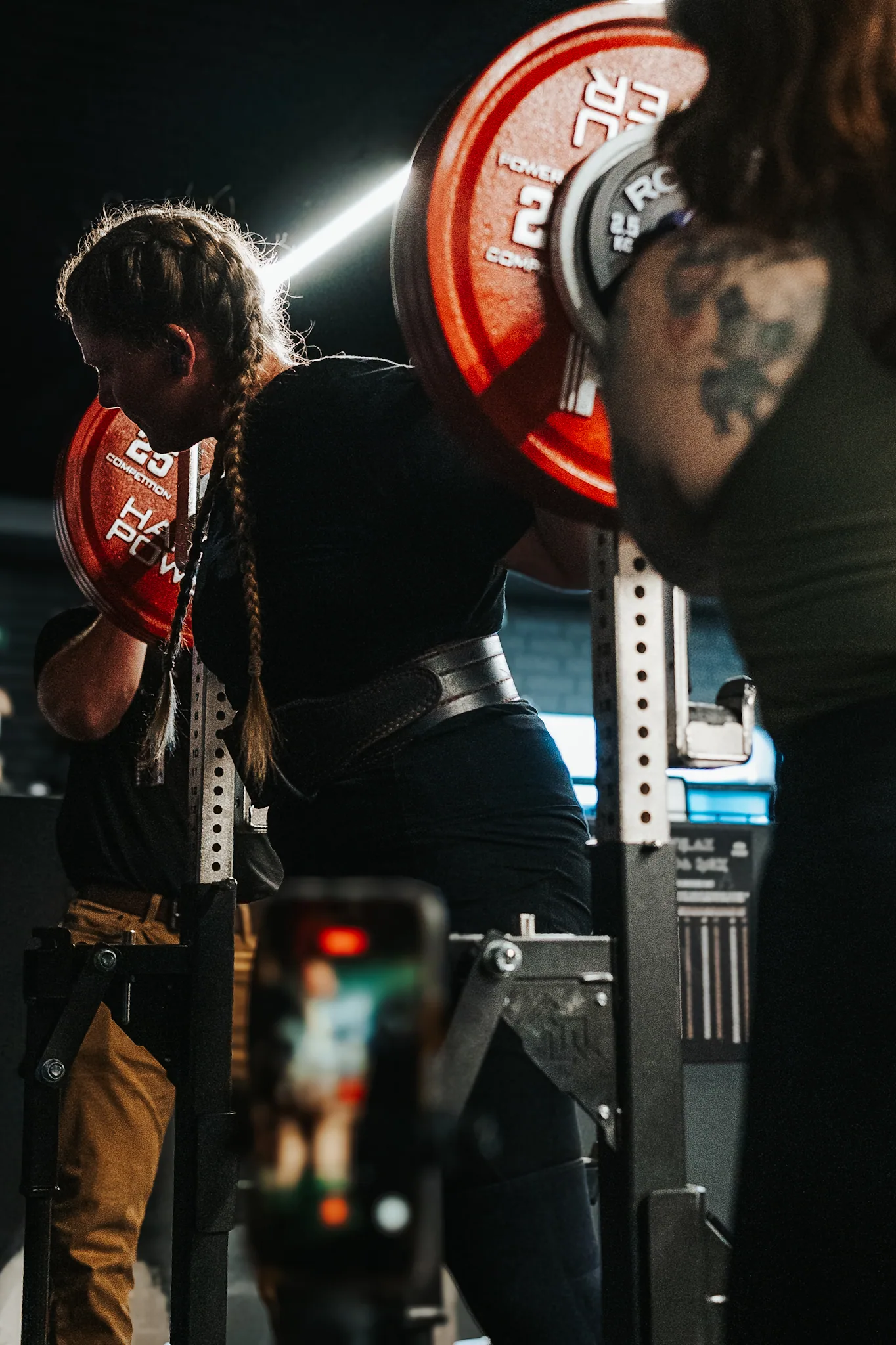 A lifter deep in a squat under red competition plates, lifting belt at the waist, a spotter either side of the rack and a phone on a tripod filming the set.