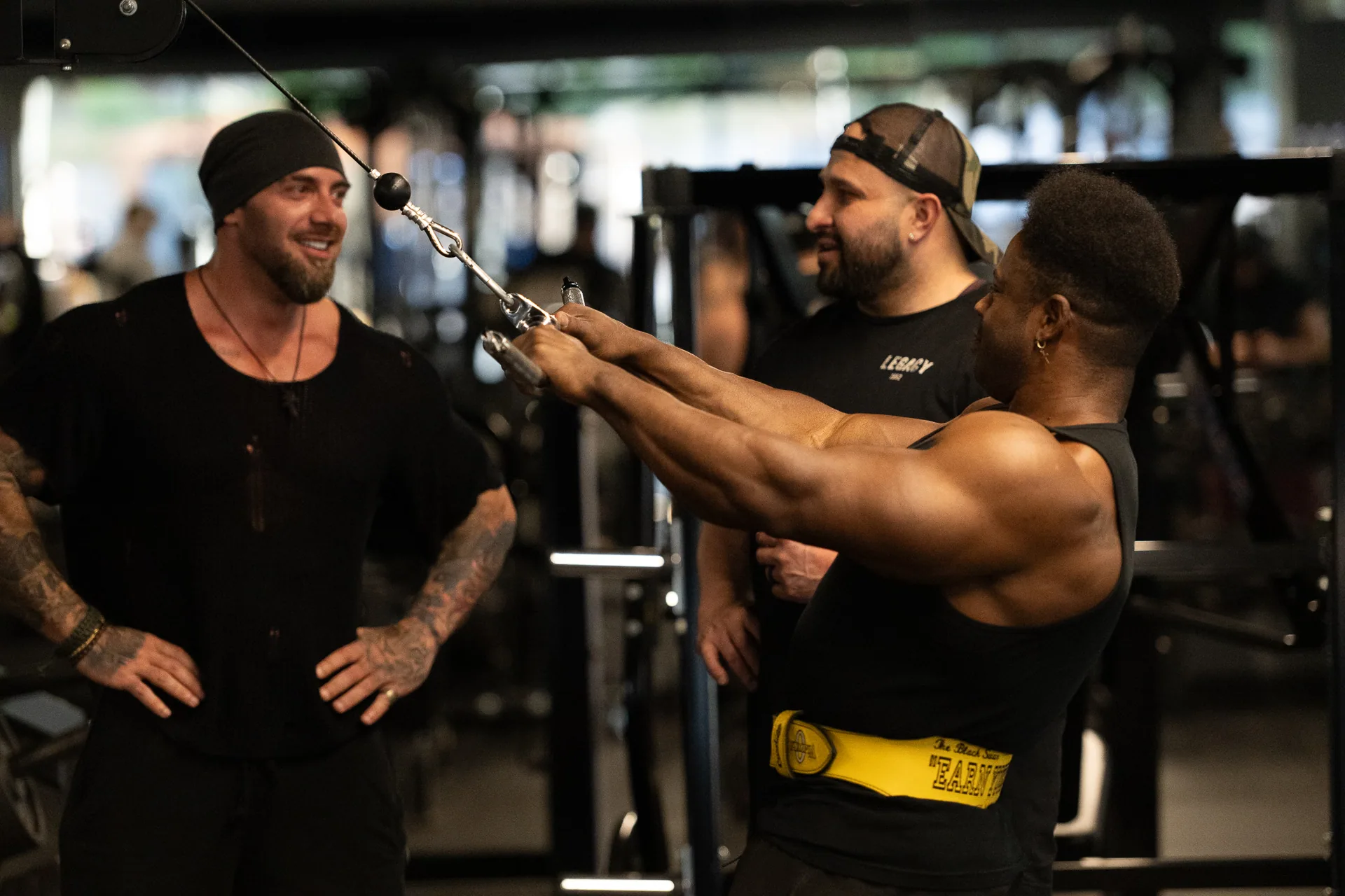 A trainer coaching two members through a cable press, all three at the same station on the main floor.