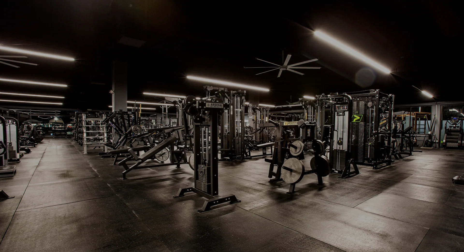 The main training floor after hours, lit low and warm, with plate-loaded machines and cable stations lined up beneath the ceiling fans.
