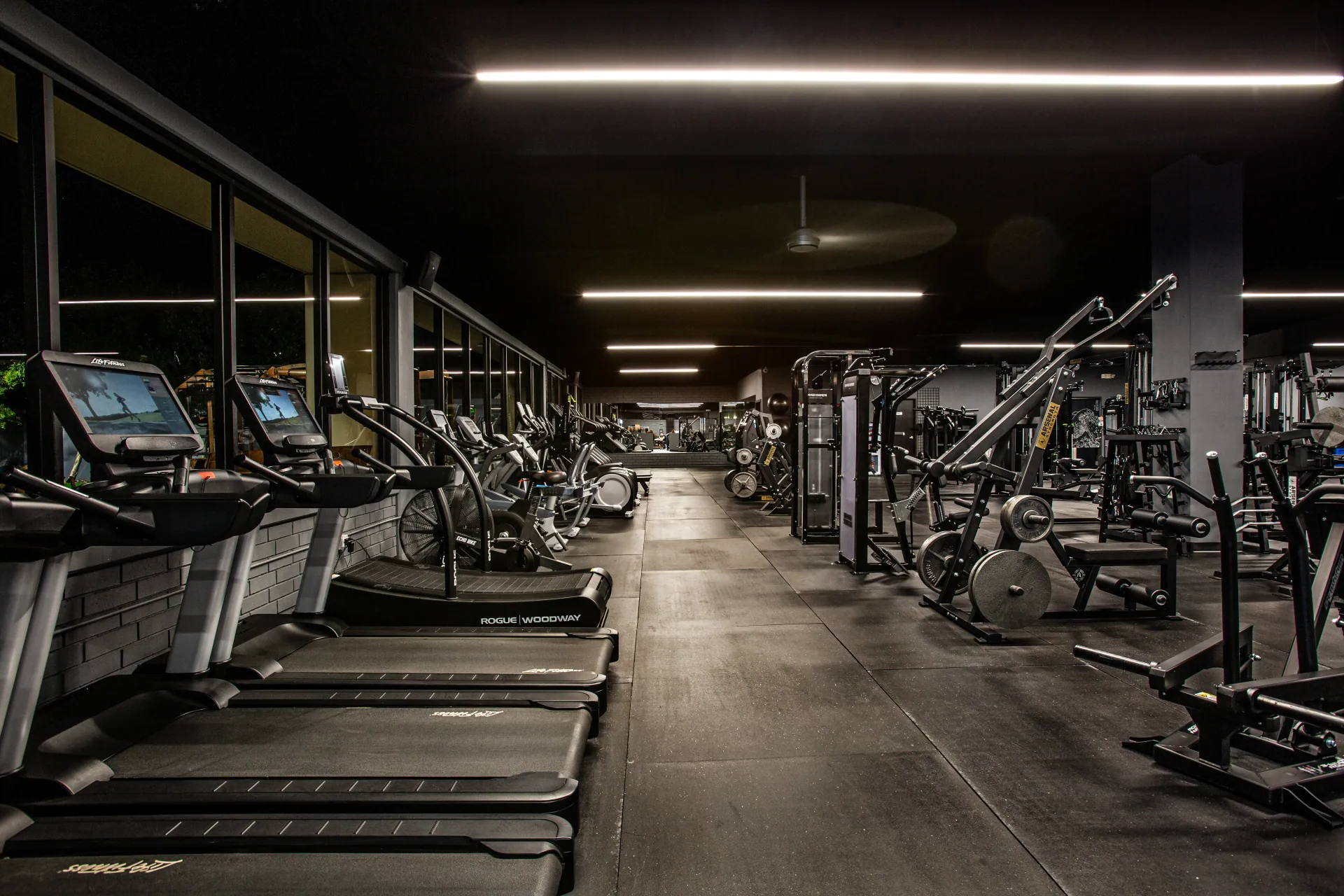 The main training floor at Athlos Iron Lair, looking down the length of the facility past the cardio row to the strength equipment