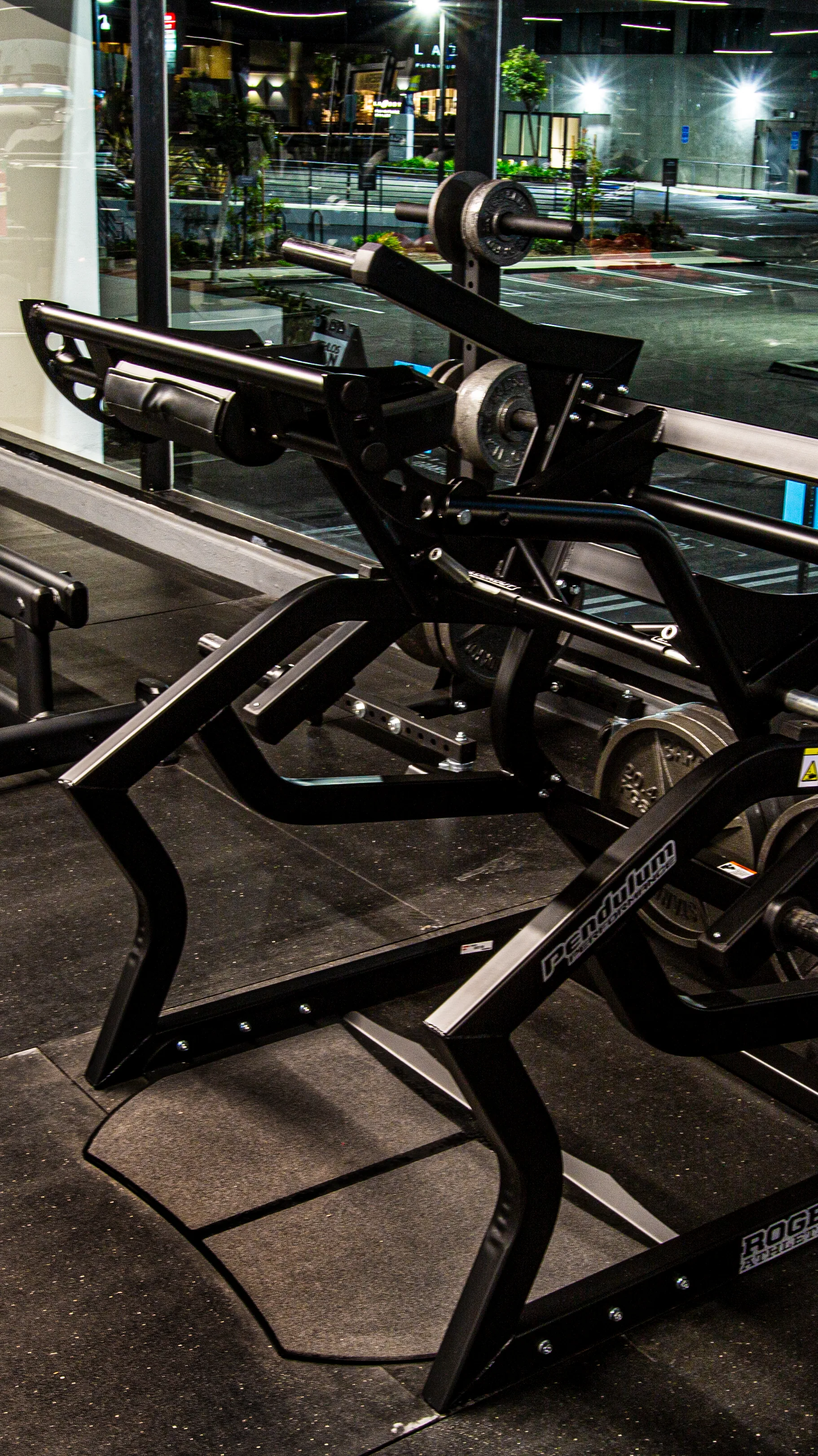 The pendulum power squat, its angled arm and shoulder pads set over the foot platform, city lights through the glass behind.