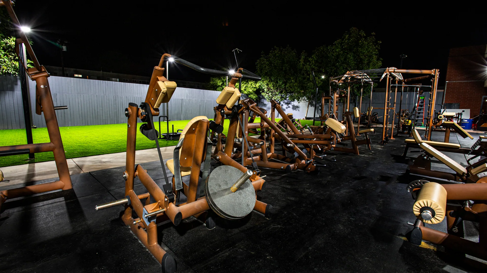 The outdoor strength zone at night, plate-loaded machines lit on the turf